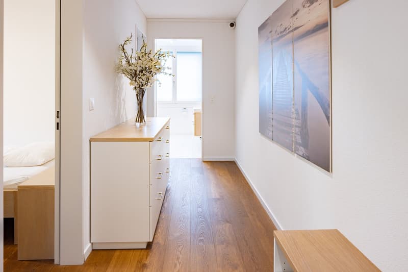 Bright and spacious hallway with hardwood floors, white walls, and a large window. There is a wooden cabinet or dresser with drawers and a vase of flowers on top. A large artwork or photograph is hung on the wall at the end of the hallway.