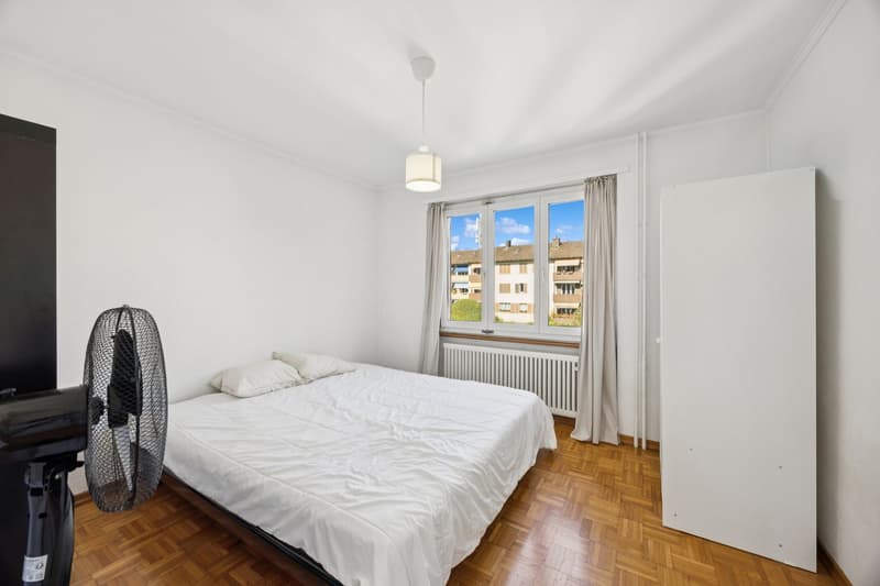 white walls, wooden floor, white sheets, two pillows, small lamp hanging from ceiling, window with curtains, building view