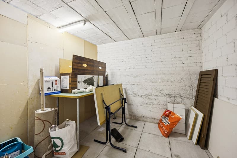 Small room, unfinished, white brick walls, tile flooring, storage boxes, desk, folding table, sacks