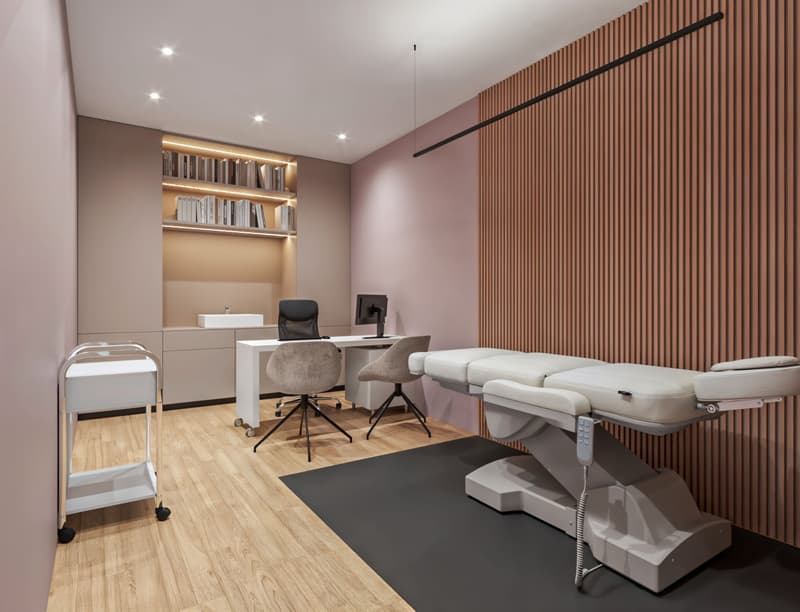 modern medical office with a desk, two chairs, a monitor, a sink, a massage table, a trolley, and a wooden wall