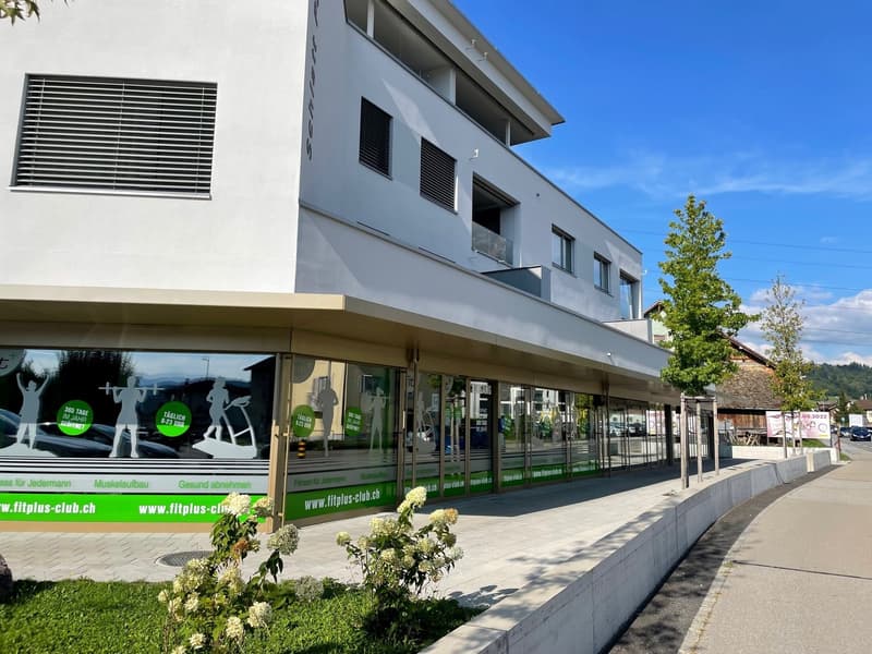 Modern white building with glass windows, green signage, and a flat roof, located on a street with a sidewalk and greenery.