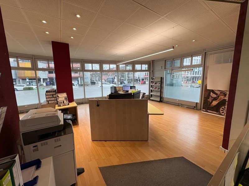 Office interior with wooden floors, glass windows, a reception desk, chairs, computers, bookshelves, large room, and an outside view.