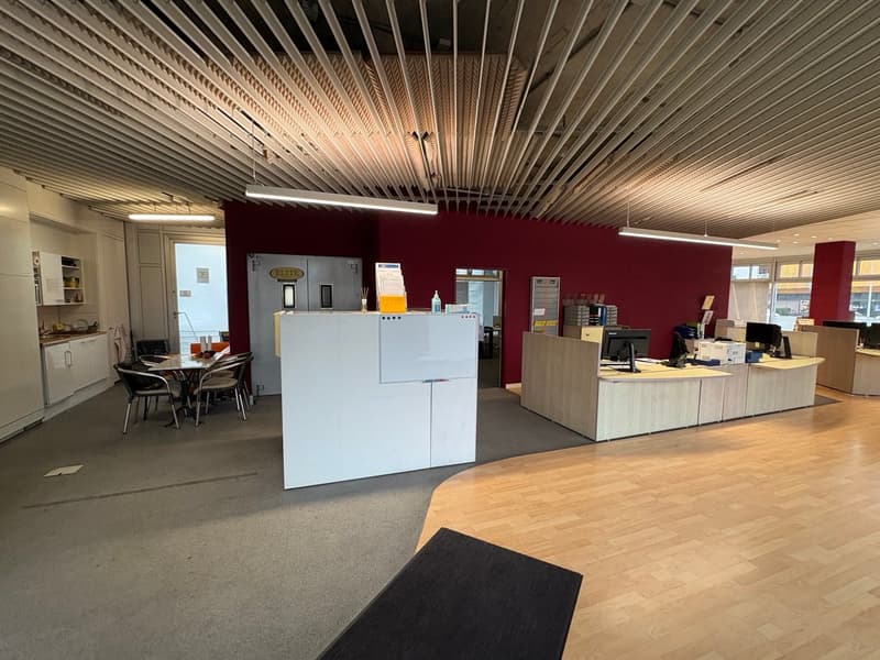 Empty office with reception area, office tables, and chairs, red wall, cabinets, white countertops, and wooden floors.