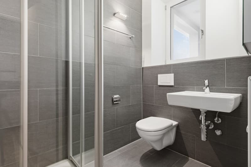 modern bathroom with gray tiles, white toilet, white sink, glass shower enclosure, wall-mounted faucet, small window