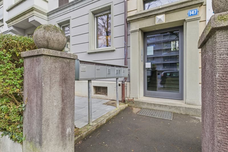 Concrete entrance with mailbox, glass door, number 147, stone pillars, paved walkway, greenery
