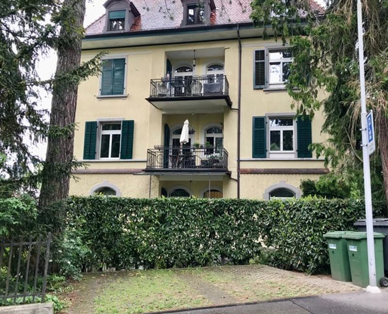 3-story yellow building with green shutters, balconies, and a tiled roof. The building is surrounded by trees and greenery.