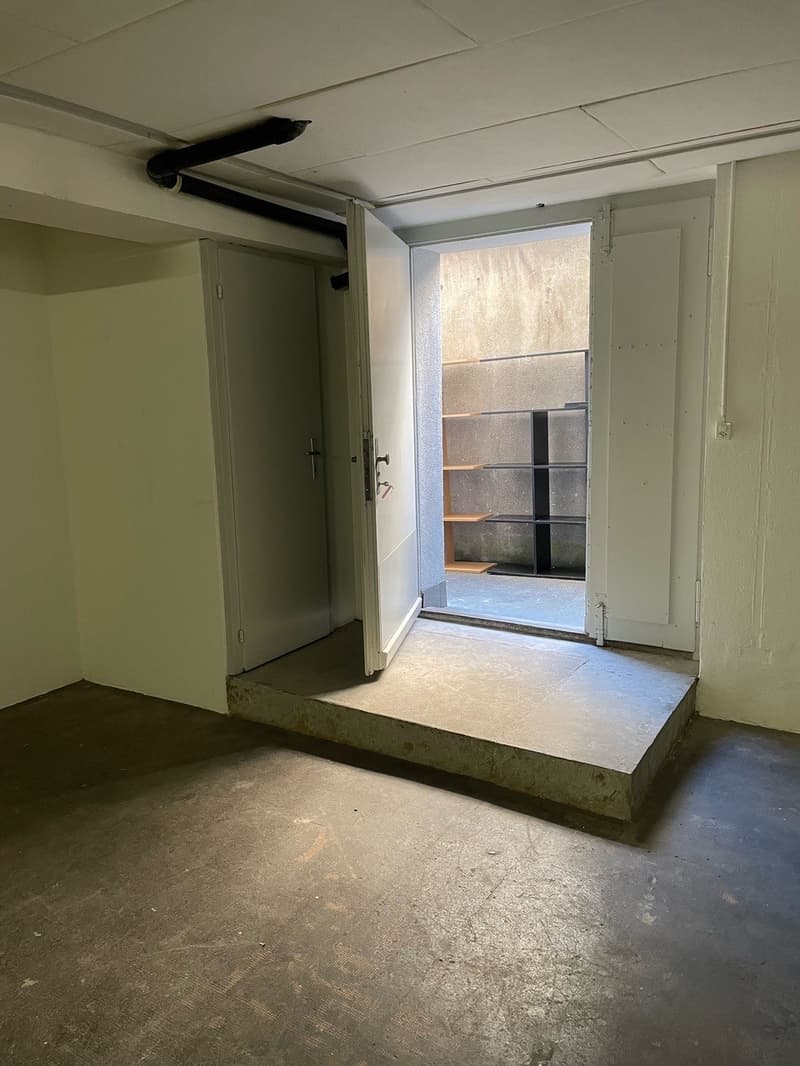The image shows an interior room with a concrete floor, white walls, and a doorway leading to an exterior space. The room appears to be in a state of renovation or construction, with exposed pipes and wiring visible on the ceiling.