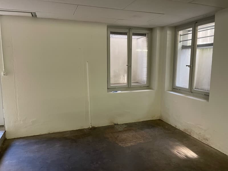 This image shows an empty room with white walls, a concrete floor, and two windows. The room appears to be in need of renovation, with some visible damage or wear on the walls and floor.