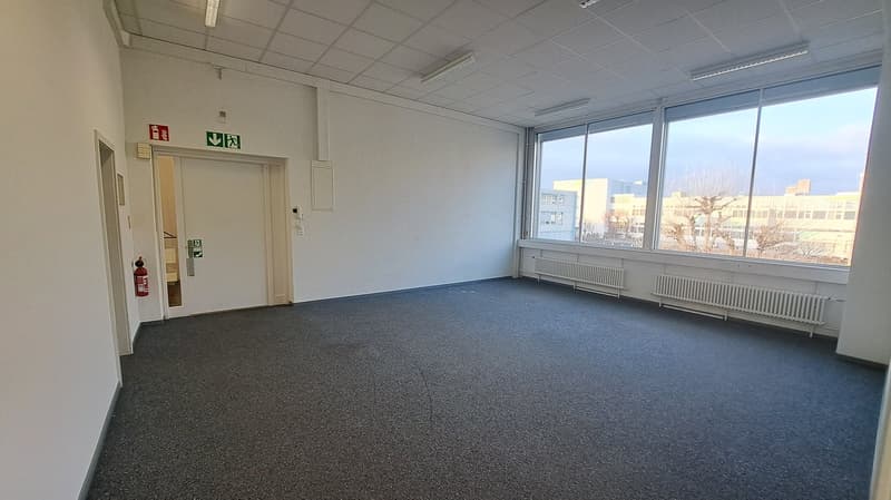 Empty room with white walls, carpet flooring, ceiling lamps, two fire extinguishers, door, glass windows