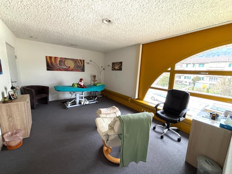 empty room with a massage table, a chair, a desk, a cabinet, a trash bin, and a window with a view of the outside