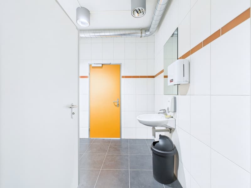 Tiled bathroom with white walls, gray floor tiles, a sink, and a yellow door. There is also a trash can and a wall-mounted appliance, likely a dishwasher, visible in the image.
