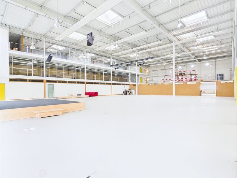 Large open warehouse space with high ceilings, exposed beams, and skylights. The space has a concrete floor and wooden platforms, as well as some furniture and equipment. The space appears to be used for storage or industrial purposes.