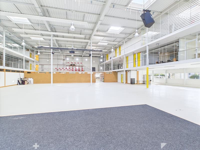 Large open space with high ceilings, concrete floor, and glass walls. The space appears to be a warehouse or industrial facility with ample lighting and storage areas.
