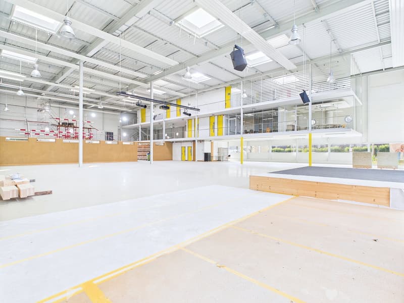 Large open warehouse space with high ceilings, white walls, and yellow accents. The space has a concrete floor with yellow markings, and there are various storage racks and equipment visible.