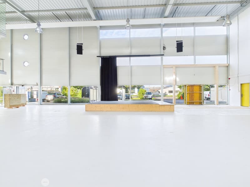 Large open commercial space with high ceilings, large windows, and a stage-like platform. The space appears to be used for events or presentations, with parking visible outside the windows.