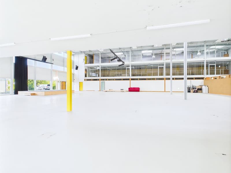 Large open commercial space with white walls, yellow support columns, and glass windows. The space appears to be empty and ready for use, with a red sofa visible in the background.