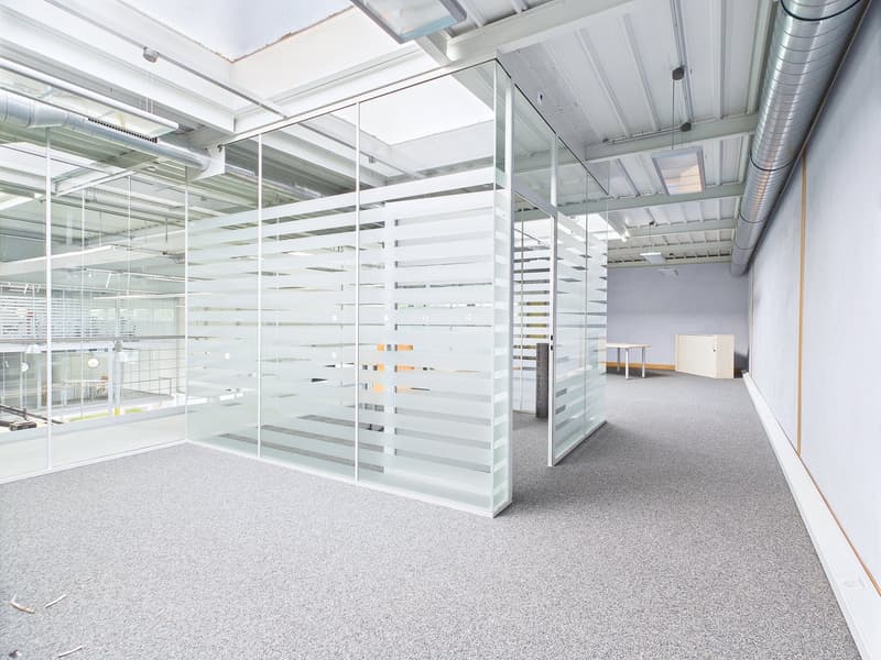Large open office space with glass partitions, concrete floors, and exposed ceiling with ventilation ducts. The space appears to be a modern, minimalist office design with ample natural light and a clean, industrial aesthetic.