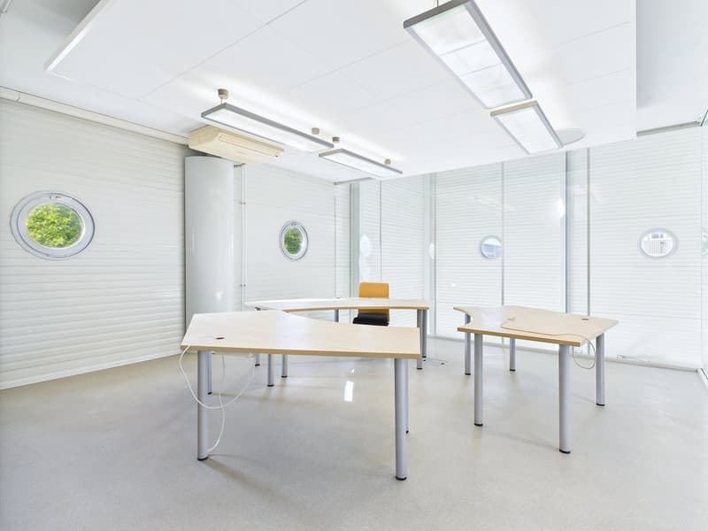 Bright and modern office space with large windows, white walls, and minimalist furniture including desks and chairs. The room is equipped with overhead lighting and appears to have a dishwasher visible.