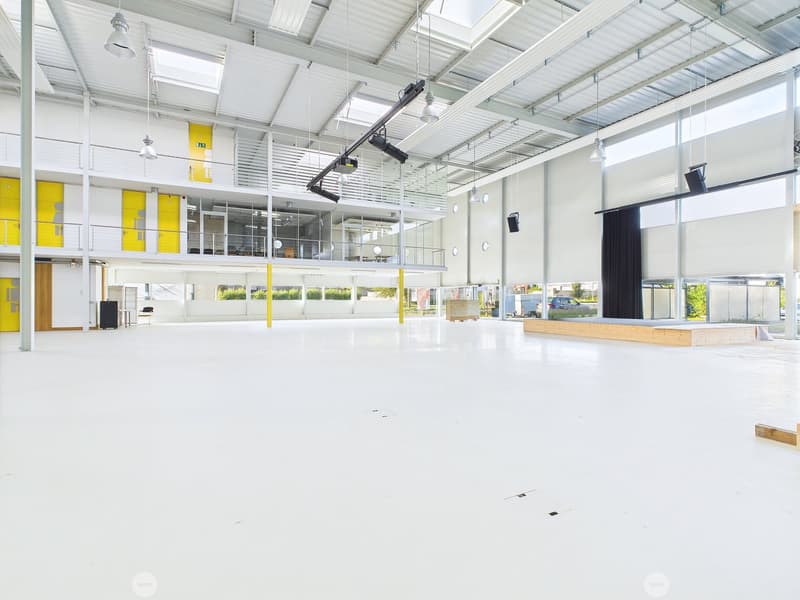 Large open commercial space with high ceilings, white walls, and large windows. The space has a stage-like area with a wooden platform and appears to be set up for events or performances. The space is well-lit with overhead lighting and has a modern, indu