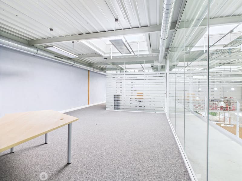 The image shows a modern, open-plan office space with a clean, minimalist design. The space features a large desk in the foreground, with glass walls and partitions separating different work areas. The ceiling is high and has exposed ductwork, giving the