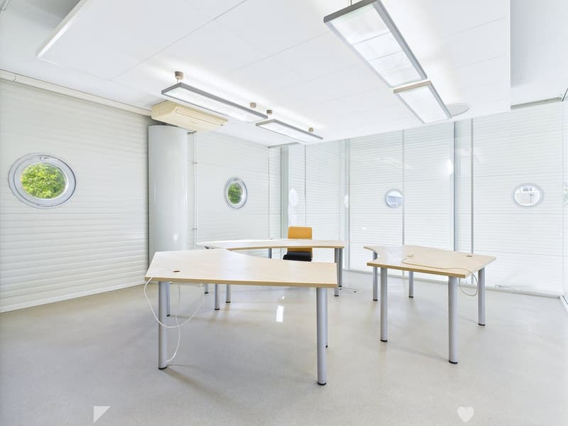Bright and modern office space with large windows, white walls, and minimalist furniture including desks and chairs. The room has recessed lighting and a clean, professional atmosphere.