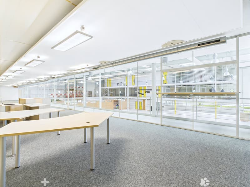 Large open office space with glass walls, desks, and shelving units. The space appears to be well-lit with overhead lighting and has a modern, minimalist design.