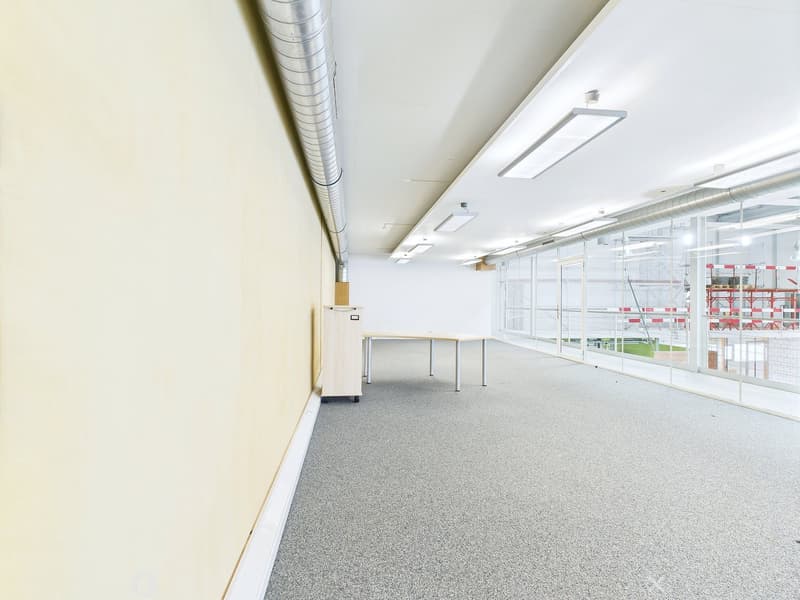 The image shows a large, open office space with white walls, a gray carpeted floor, and overhead lighting fixtures. There is a table visible in the foreground, and the space appears to have large windows along one side, providing natural light. The overal