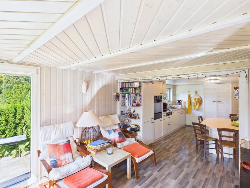 white ceiling, wooden wall, armchair, side table, shelf with books, kitchen, dining table and chairs, glass door, garden view
