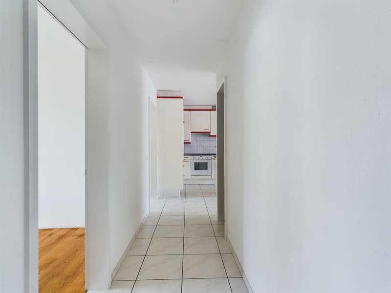Empty hallway with white tiles, white walls, wooden floor in one part, kitchen in the background