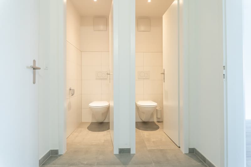 Two toilets, white walls, tiled floor, white doors, light fixtures