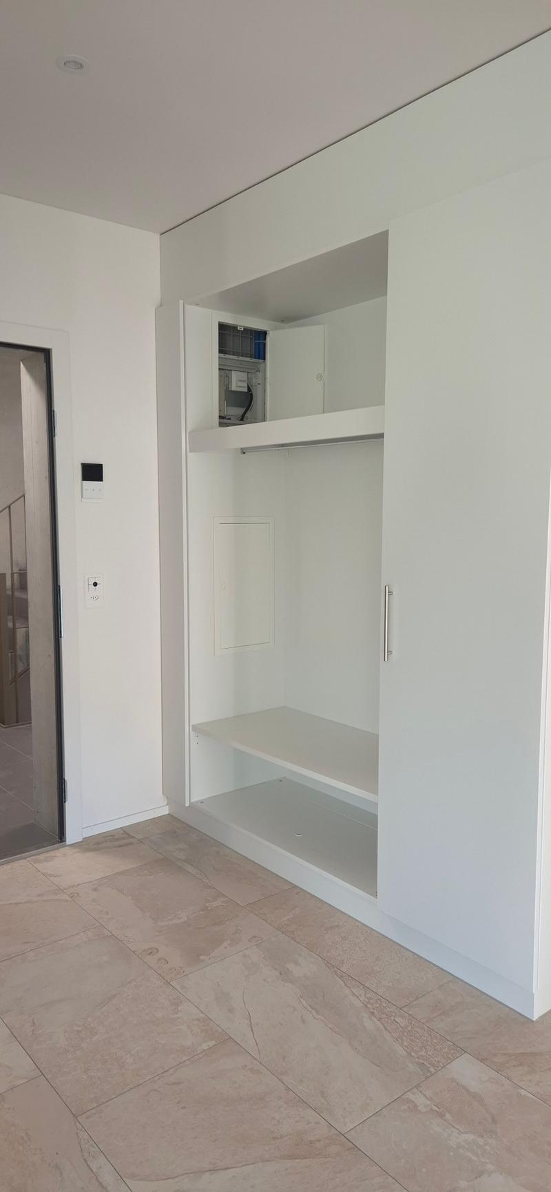 Empty room with white walls, tiled floor, built-in wardrobe with shelves, and an open door leading to another room.