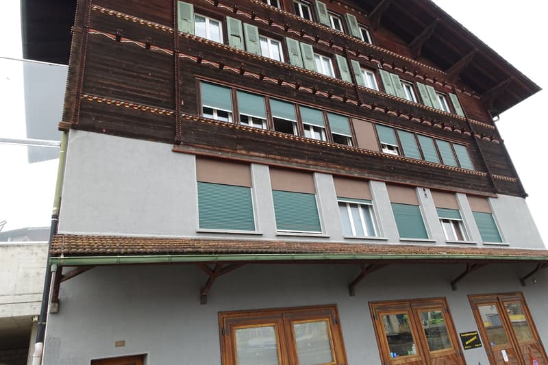 3-story traditional Swiss chalet-style building with wooden exterior, green shutters, and a balcony on the upper floor