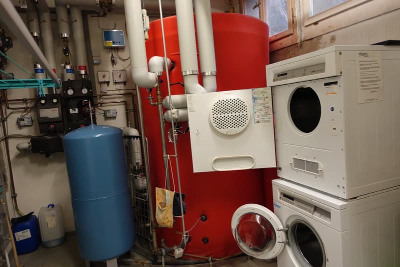 The image shows a utility room with various appliances and equipment. There is a large red water heater, a washing machine, a dryer, and other plumbing and electrical equipment. The room appears to be used for storage and housing the home's utilities.