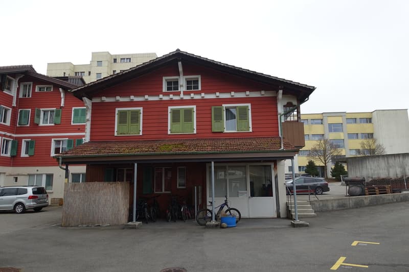 2-story red wooden building with green shutters, parking spaces in front, surrounded by modern apartment buildings