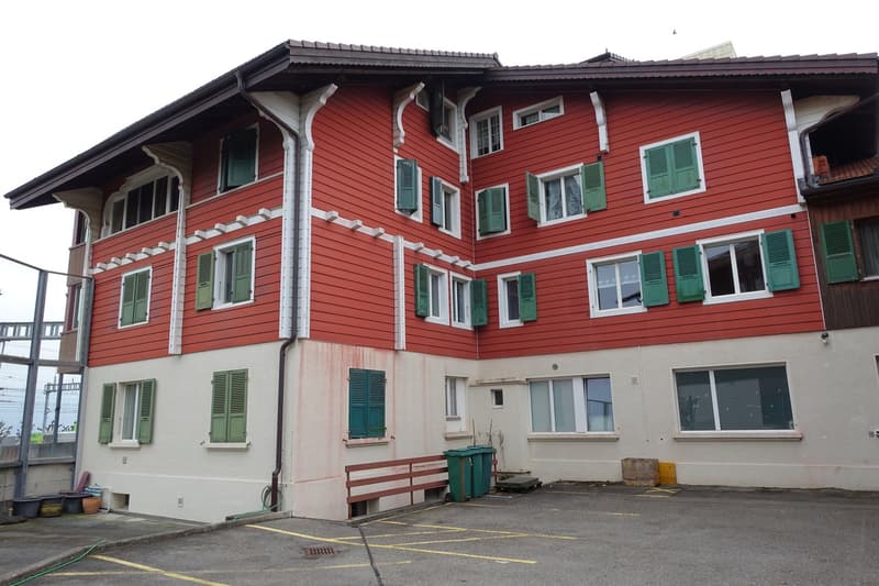 A multi-story red wooden building with green shutters and a parking area in front