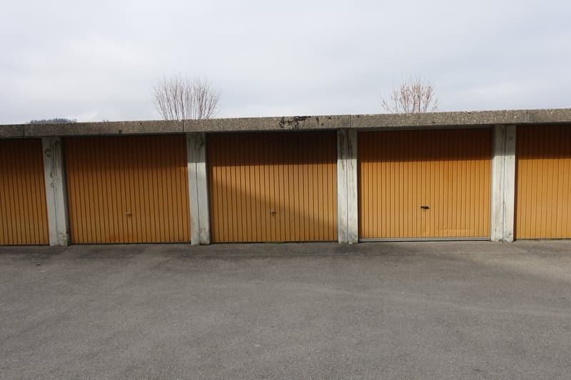 The image shows a row of three garage doors made of wooden panels in an orange-brown color. The doors are set into a concrete or stone structure with a flat roof. The ground in front of the garages is paved with asphalt or a similar material.
