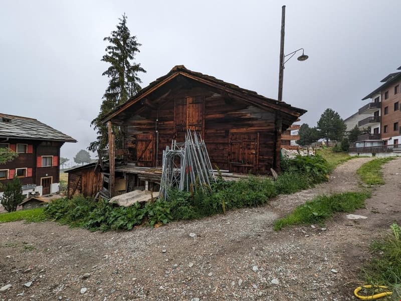 wooden building on slope, two smaller houses, greenery around, dirt path in front
