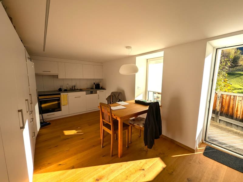 The image shows an open-plan kitchen and dining area with a wooden dining table and chairs, a kitchen with white cabinets and appliances, and a large window overlooking a balcony or terrace area.