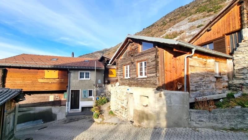 Traditional alpine-style wooden and stone houses, with slanted roofs and balconies. The buildings appear to be located in a mountainous, rural area with a scenic backdrop.
