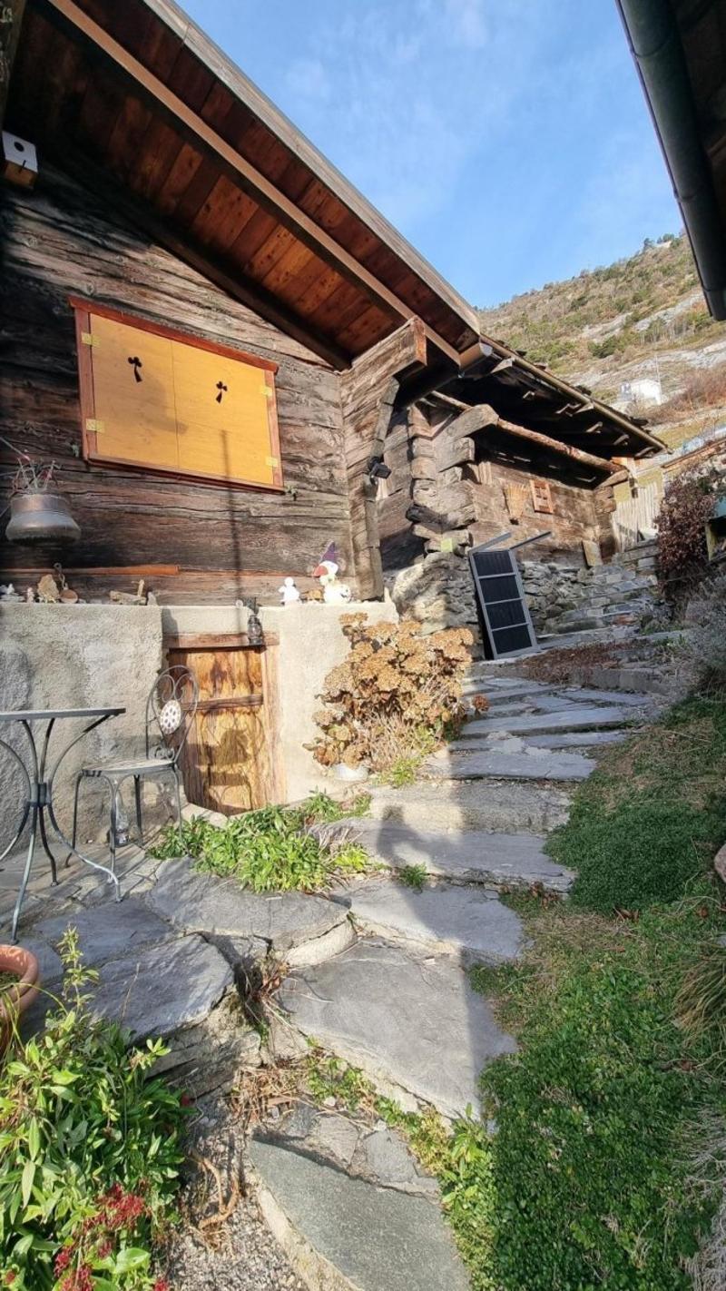 Rustic wooden cabin with a slanted roof, stone steps leading up to the entrance, and a small patio or balcony area. There appears to be a fireplace visible on the exterior of the cabin.