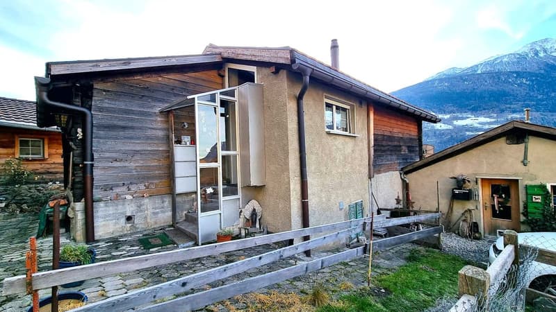 Two-story wooden house with a raised ground floor, stone exterior, and a balcony or terrace. The house is surrounded by a wooden fence and has a small yard with some outdoor items.