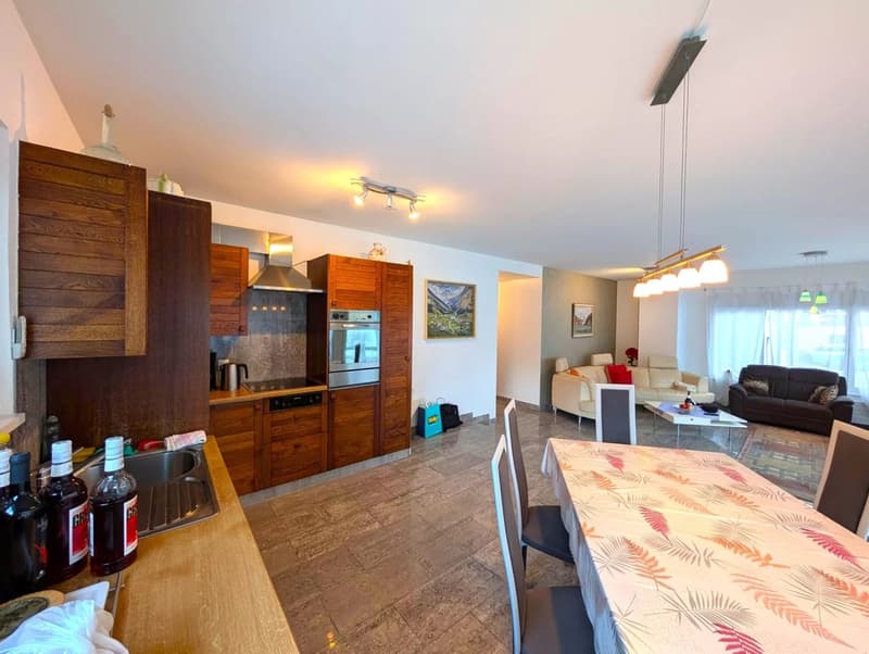 The image shows an open-concept living space with a kitchen, dining area, and living room. The kitchen features wooden cabinets and stainless steel appliances, including a dishwasher. The dining area has a patterned table and chairs, while the living room