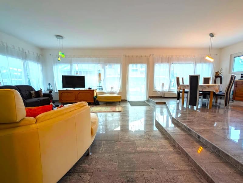 Spacious open-plan living area with large windows, hardwood floors, yellow and black sofas, TV stand, dining table and chairs, and a raised platform with glass doors leading to a balcony or terrace.