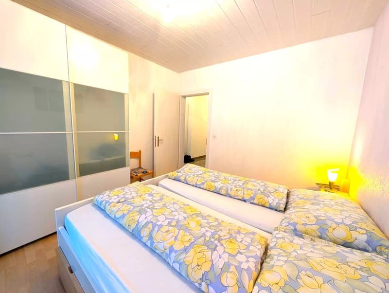 Bedroom with two beds, each with floral bedspreads, white walls, white ceiling, open door, bedside table with lamp, sliding wardrobes, wooden chair