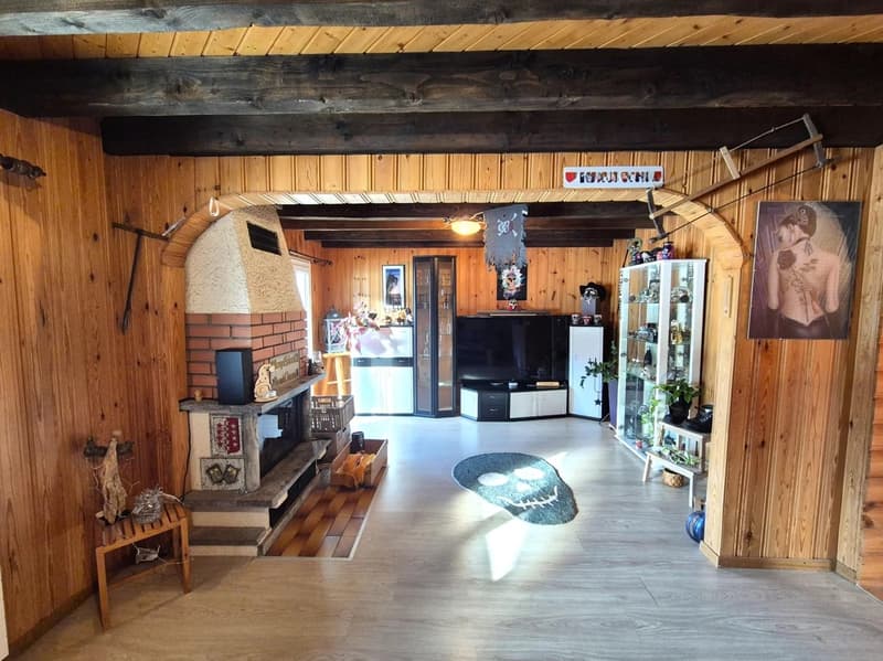 Rustic wooden interior with exposed beams, fireplace, TV stand, shelves with decorative items, and a large area rug on the floor.