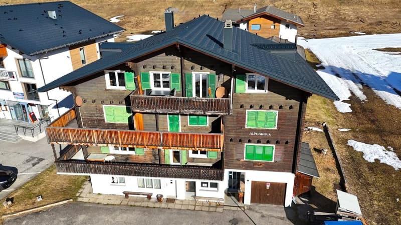 3-story alpine-style building with wooden balconies, green shutters, and a black tiled roof. The building has the name 'Alpenblick' written on it.