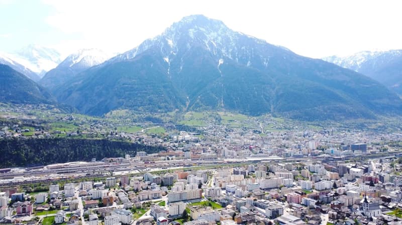 Aerial view of a mountainous city with a large snow-capped peak in the background, surrounded by residential and commercial buildings, railways, and green spaces.