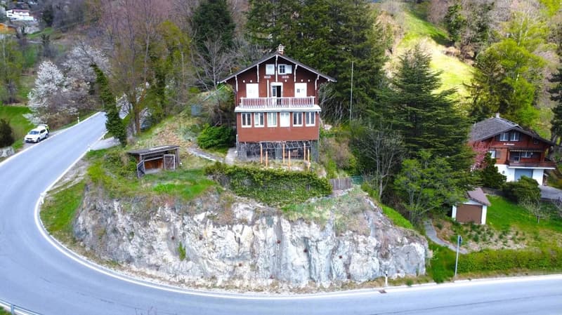 A two-story wooden chalet-style house situated on a hillside, surrounded by lush greenery and trees. The house has a raised ground floor and a balcony overlooking the scenic landscape. There is a road winding around the property and another smaller buildi