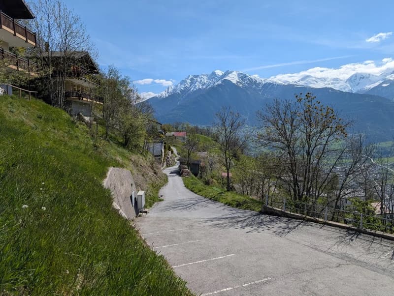 The image shows a scenic mountain landscape with snow-capped peaks in the background. In the foreground, there is a winding path or road surrounded by lush greenery, including trees and grass. There are also some buildings or structures visible on the hil