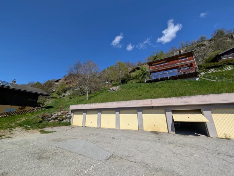 A multi-story wooden building with a balcony, surrounded by a grassy hill and trees. There is a row of garages or storage units in the foreground.